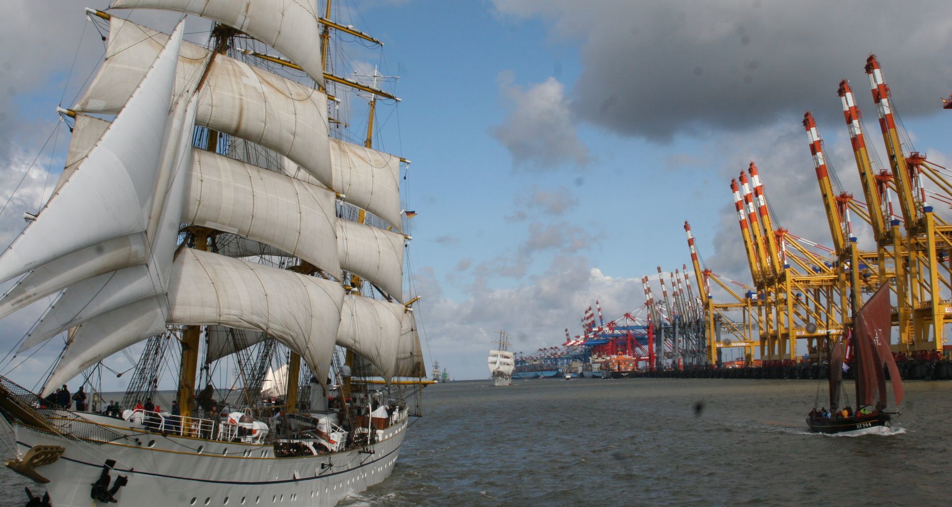 A sailing ship on a cruise, in the background a container terminal.