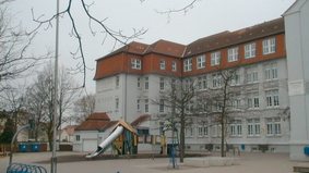 Auf dem Bild ist ein Pausenhof mit einigen kahlen Bäumen und einem Spielgerüst abgebildet. Im Hintergrund sieht man die Astrid-Lindgren-Schule.