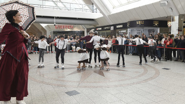 Kinder und eine erwachsene Person in historischen Kostümen unter einer gläsernen Kuppel in einem Einkaufszentrum. Alle Personen tanzen mit Rollschuhen. Eine Gruppe Personen steht am Rand und schaut zu.