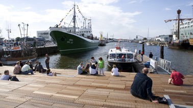 Visitors sit on a wooden deck and look out over a harbor basin.