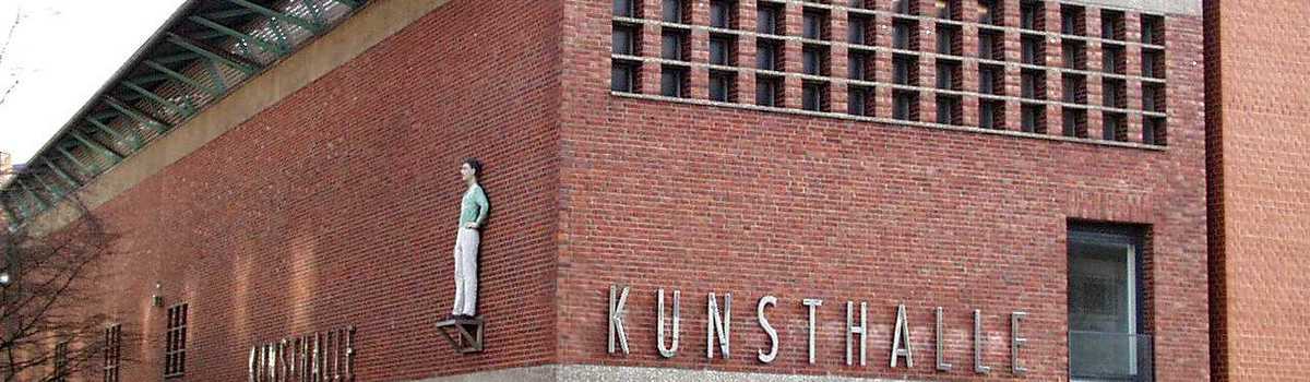 Außenaufnahme einer Kunsthalle. An der Fassade ist eine Figur aufgehängt. 