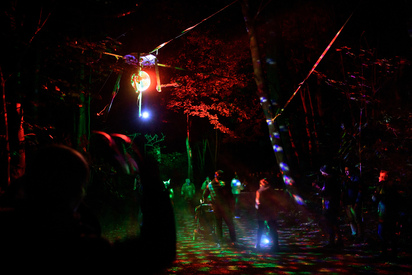 Fußweg im Park mit großer Discokugel, die über dem Weg hängt. Bunte Strahlen beleuchten die Besucher und den Wald.