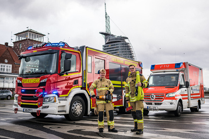Ein HLF und ein RTW der Feuerwehr Bremerhaven vor der Skyline Bremerhavens. Im Vordergrund zwei Einsatzkräfte.