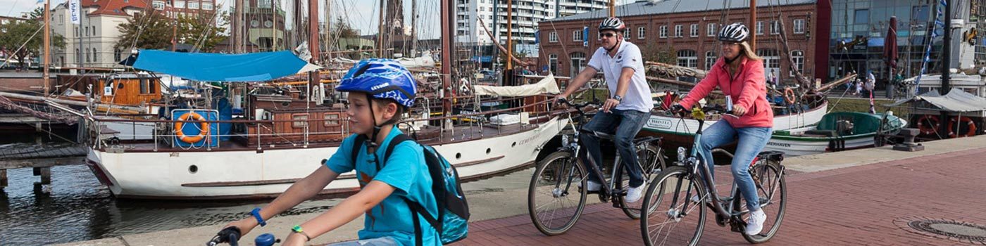Eine Familie fährt mit Fahrrädern an Schiffen vorbei.
