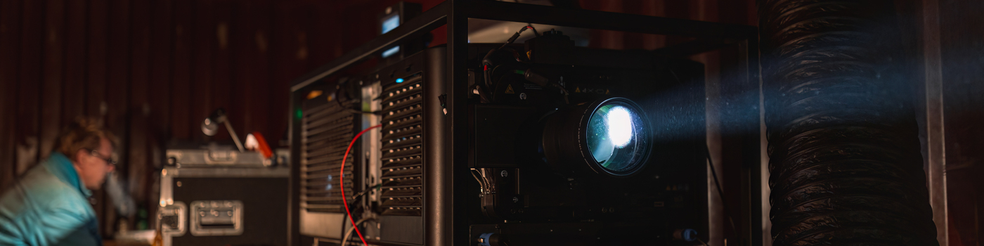 Leuchtende Projektorlinse und eine Vielzahl an Projektionstechnik in einem dunklen Containerturm. 
