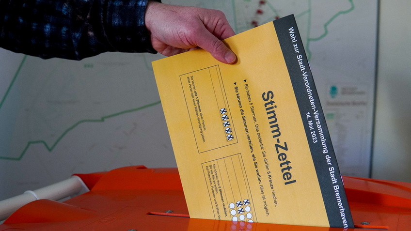 Hand steckt einen gelben Wahlzettel in eine hellrote Urne.