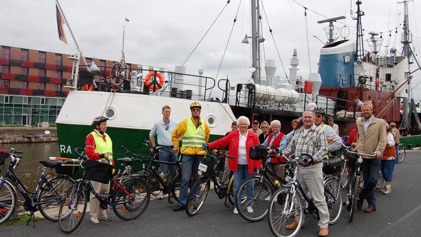 Ein Gruppe Radfahrer steht vor einem Museumsschiff.