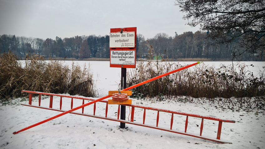 Uferkante eines zugefrorenen Sees. Schnee liegt an Land und auf dem Eis. Im Vordergrund ein Schild: Betreten des Eises verboten. An dem Schild sind Rettungsgeräte zur Eisrettung (Leiter, Stange mit Haken und Leine)