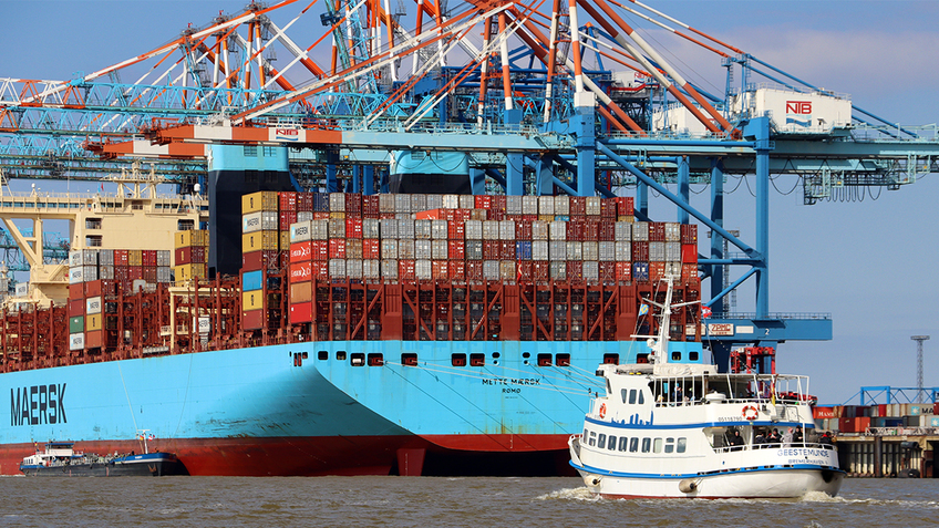 A ship is driving on a river, container terminal in the background.