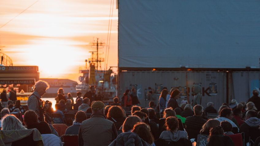 Menschen sitzen bei Sonnenuntergang im Schaufenster Fischereihafen und warten auf den Beginn des Kino im Hafens 2022.