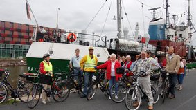 A group of cyclists is facing a museum ship.