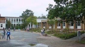 Auf dem Pausenhof spielen einige Kinder. Im Hintergrund ist die Gaußschule I zu sehen. In den Beeten vor der Schule stehen mehrere Bäume.
