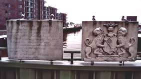 Zwei Gedenktafeln aus Stein, im Hintergrund ein Hafenbereich.