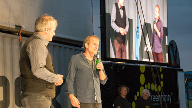 Zwei Männer stehen auf der Bühne vor dem Containerturm und halten Mikrofone in der Hand. Im Hintergrund werden sie nochmal auf einem Bildschirm per Kamera übertragen.