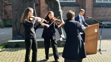 Ein Mann steht vor einer lebensgroßen Skulptur an einem Rednerpult. Zwei Frauen spielen daneben Violine. Eine weitere Frau hält einen Notenständer fest.