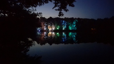 Ein See bei Nacht. Auf dem gegenüberliegenden Seeufer werden die Bäume in verschiedenen Farben beleuchtet. 