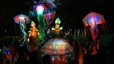 Kostümierte und leuchtende Gestalten wandelnde bei Dunkelheit durch eine Menschenmenge. Die Kostüme sind bunt und leuchten. 