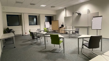 Seminarraum mit 6 dreieckigen Einzeltischen in U-Form vor einer interaktiven Tafel, zwei Stellwänden und einem Flipchart.