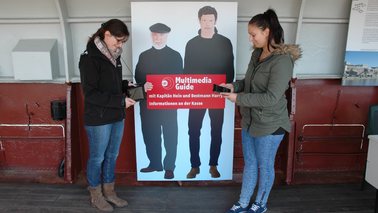 Zwei junge Frauen stehen vor einem Plakat und halten ein Hand in der Hand.