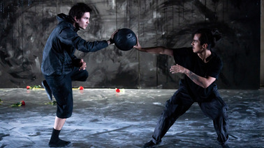 Zwei Männer stehe nebeneinander und gucken sich an. Sie halten beide gemeinsamen einen Ball mit ihren Händen zwischen sich. Das Bild und ihre Kleidung ist dunkel gehalten.