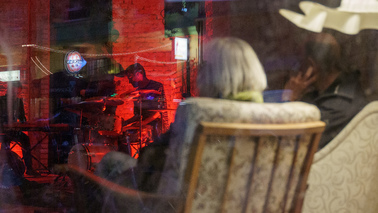 Durch eine Fensterscheibe ist ist eine in einem Sessel sitzende Dame fotografiert. Im Hintergrund spielt ein Mensch Gitarre.