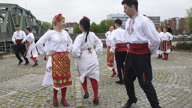 Menschen in traditioneller bulgarischer Kleidung (weiß und rot) tanzen einen Volkstanz auf einem gepflasterten Platz