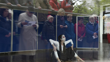 Eine weiß geschminkte Frau hinter einer Glasscheibe. In der Scheibe spiegeln sich die Zusehenden davor.