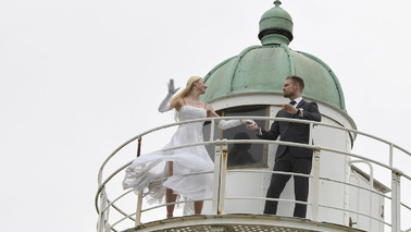 Ein Paar tanzt auf der Reling eines Leuchtturms. Der Leuchtturm und das Kleid der Frau sind weiß. Die Kuppel des Leuchtturms ist kupfergrün. Der Mann trägt einen dunklen Anzug.