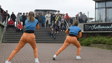 Zwei Mädchen tanzen HipHop. Sie tragen die gleichen orangen Hosen und blauen Oberteile. Sie tanzen draußen, hinter ihnen steht eine Gruppe Personen und schaut zu.