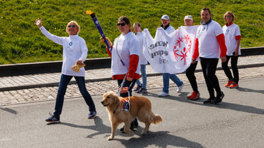 7 Personen in Special Olympics Fackellauf T-Schirts laufen auf einer Straße. 3 Personen tragen eine Special Olympics Banner. Eine weitere Person trägt die brennde Fackel. Sie wird von ihren Blindenführhund gegleitet.