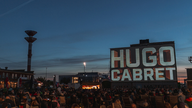 Dämmerung im Schaufenster Fischereihafen. Viele Menschen sitzen vor der Leinwand mit der Titelprojektion vom Film "Hugo Cabret". 