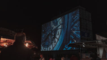 Schaufenster Fischereihafen bei Nacht. Filmleinwand ist leicht von der Seite zu sehen. Ein Mann steht davor, dahinter erkennt man Teile des Publikums, welches den Film "Hugo Cabret" schaut. 