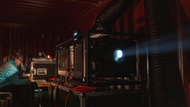 In einem dunkeln Raum steht ein großer Filmprojektor. Im Hintergrund arbeitet eine Person an einem Computer.