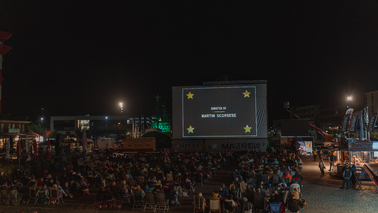 Schaufenster Fischereihafen bei Nacht. Viele Menschen sitzen vor der erleuchteten Filmleinwand und schauen den Film "Hugo Cabret".