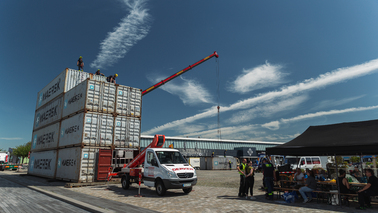 Vor dem fast fertigen Containerturm steht ein Hebefahrzeug der Firma Gerken. Im Hintergrund wird ein weiterer Container am Kran befestigt. 
